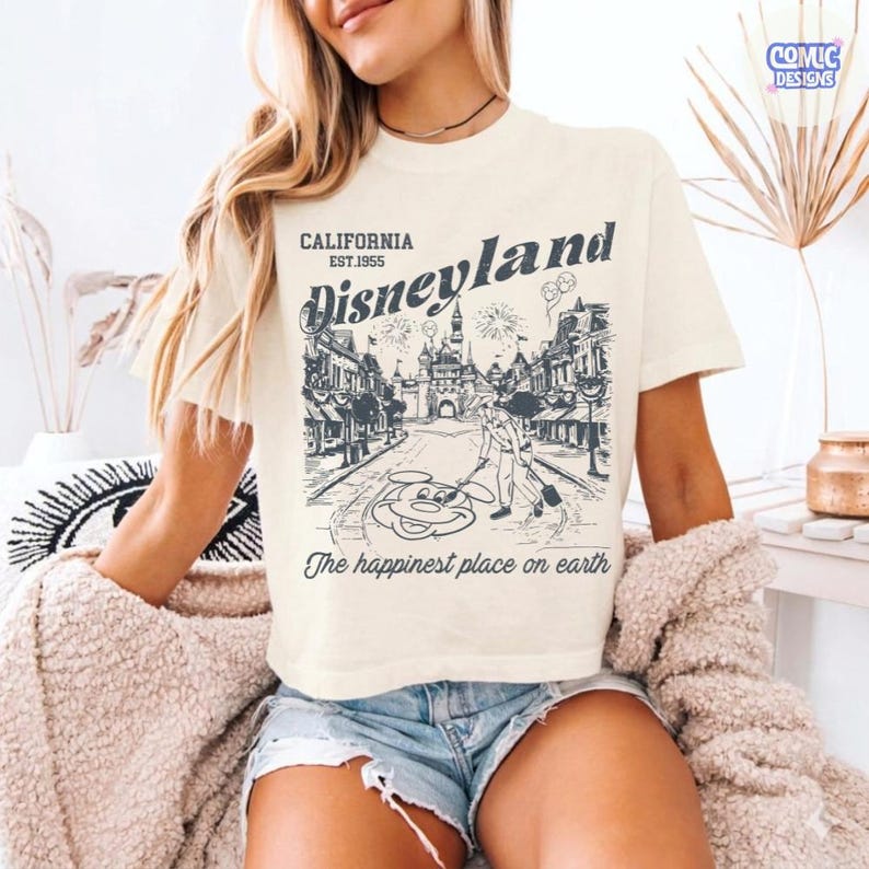 May include: Cream-coloured t-shirt with a vintage Disneyland design. The graphic includes the text "CALIFORNIA EST.1955 Disneyland" with an illustration of Main Street, U.S.A., and Mickey Mouse. The phrase "The happiest place on earth" is printed below.
