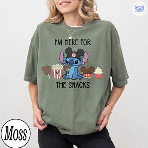 May include: Sage green t-shirt featuring a blue cartoon character wearing a Mickey Mouse hat, surrounded by snack illustrations. The text "I'M HERE FOR THE SNACKS" is printed above and below the character.