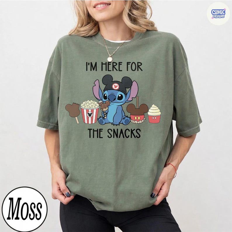 May include: Sage green t-shirt featuring a blue cartoon character wearing a Mickey Mouse hat, surrounded by snack illustrations. The text "I'M HERE FOR THE SNACKS" is printed above and below the character.