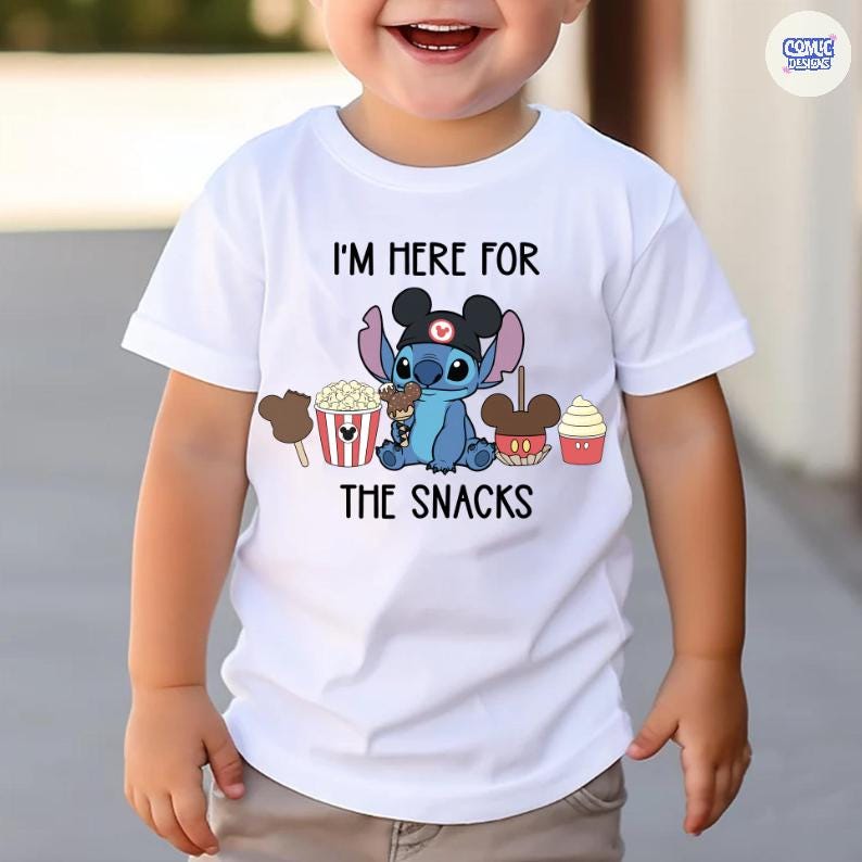 May include: White t-shirt with a graphic of the character Stitch wearing a Mickey Mouse hat and holding ice cream. The text "I'M HERE FOR THE SNACKS" is printed above and below the character. The shirt also features images of popcorn, ice cream, and cupcakes.