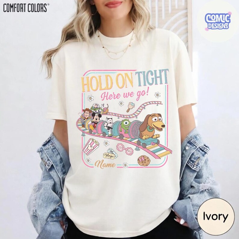 May include: Ivory-colored t-shirt with a colorful graphic featuring a cartoon train with characters and the text "HOLD ON TIGHT Here we go!". The shirt also has the text "Name" and "COMIC DESIGNS".