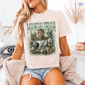 May include: Cream-colored t-shirt featuring the word "MANDALORIAN" in green block letters above a graphic of characters from the Star Wars series. The graphic includes images of the Mandalorian and other characters in helmets and armor.