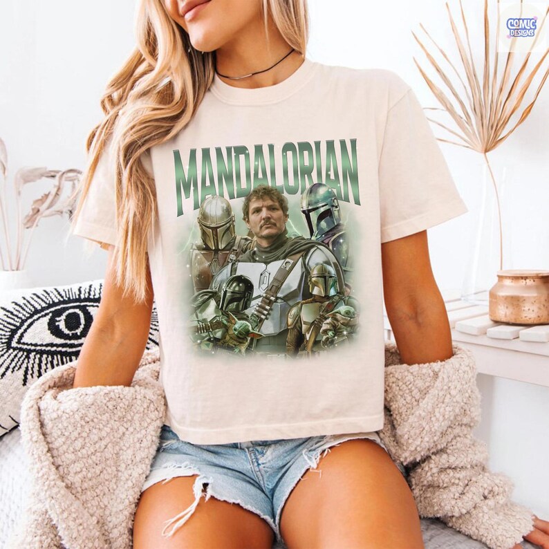 May include: Cream-colored t-shirt featuring the word "MANDALORIAN" in green block letters above a graphic of characters from the Star Wars series. The graphic includes images of the Mandalorian and other characters in helmets and armor.