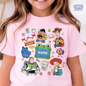 May include: A light pink t-shirt featuring a colorful graphic print of various animated characters including a space ranger, a cowgirl, a cowboy, a potato figure, and a spork. The design includes a green frog-shaped frame with the text Name.