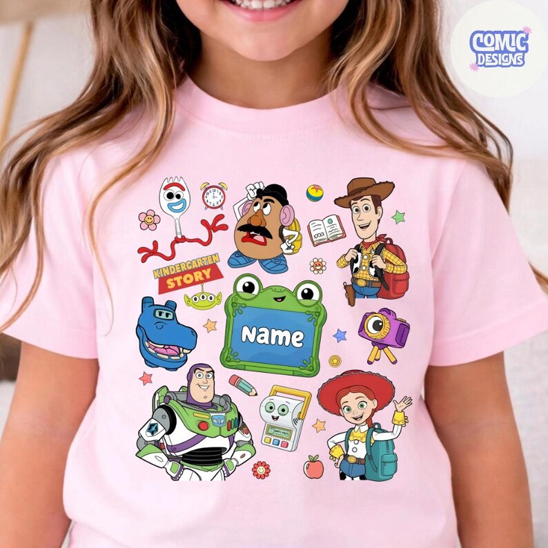 May include: A light pink t-shirt featuring a colorful graphic print of various animated characters including a space ranger, a cowgirl, a cowboy, a potato figure, and a spork. The design includes a green frog-shaped frame with the text Name.
