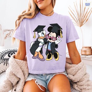 Puede incluir: Camiseta lila claro con un gr&aacute;fico de Daisy Duck y Minnie Mouse con atuendos de graduaci&oacute;n. Daisy Duck y Minnie Mouse llevan vestidos y gorras de graduaci&oacute;n negros con borlas doradas. Minnie Mouse tiene gafas rosas.