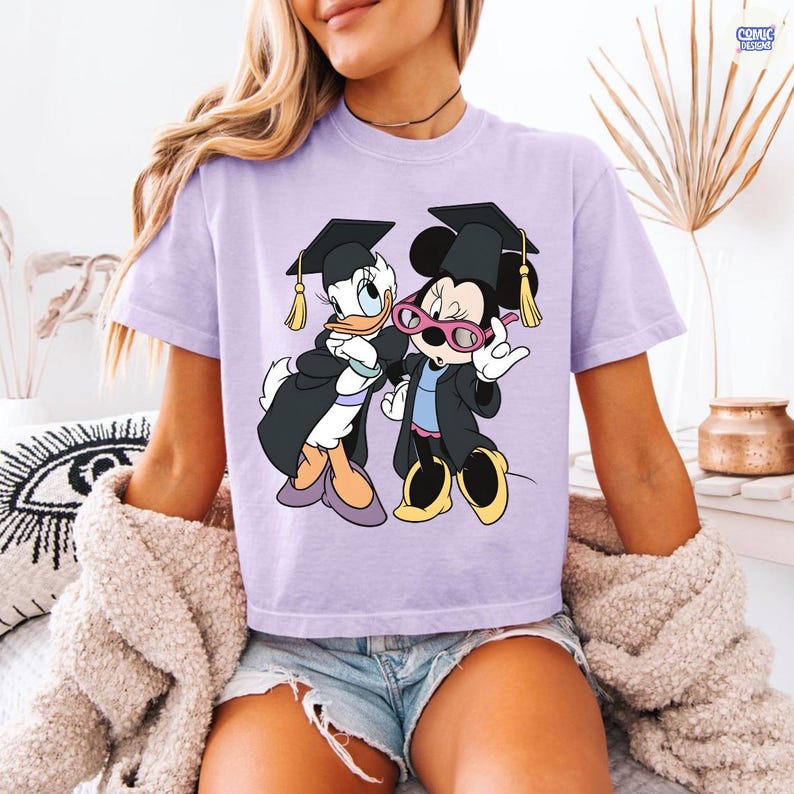 Puede incluir: Camiseta lila claro con un gr&aacute;fico de Daisy Duck y Minnie Mouse con atuendos de graduaci&oacute;n. Daisy Duck y Minnie Mouse llevan vestidos y gorras de graduaci&oacute;n negros con borlas doradas. Minnie Mouse tiene gafas rosas.