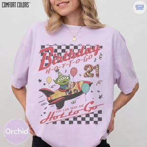 May include: Orchid-colored t-shirt with a graphic featuring a cartoon alien in a rocket ship, balloons, and the text "Birthday Hot to Go 21". The shirt has a vintage, distressed design.