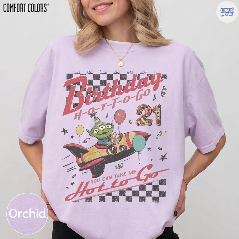 May include: Orchid-colored t-shirt with a graphic featuring a cartoon alien in a rocket ship, balloons, and the text "Birthday Hot to Go 21". The shirt has a vintage, distressed design.