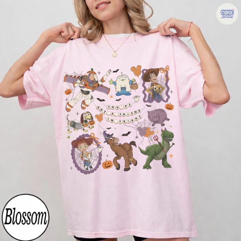 May include: A light pink t-shirt featuring a graphic print of various animated characters in Halloween costumes, including a space ranger, a cowboy, and a dinosaur. The text "YOU'VE GOT A FRIEND IN FRIGHT" is centered among spiderwebs and pumpkins.