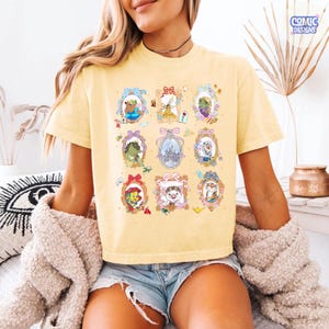 Puede incluir: Una camiseta amarillo claro con una cuadr&iacute;cula de retratos de personajes de dibujos animados en marcos ornamentados. Los marcos tienen lazos y elementos decorativos. La camiseta tiene el texto "COMIC DESIGNS" en la esquina superior derecha.