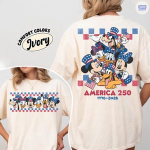 May include: Ivory-colored t-shirt featuring a graphic of Disney characters in patriotic attire, including Mickey and Minnie Mouse, Donald and Daisy Duck, and Pluto. The shirt has the text "AMERICA 250 1776-2026" and a Comfort Colors label.