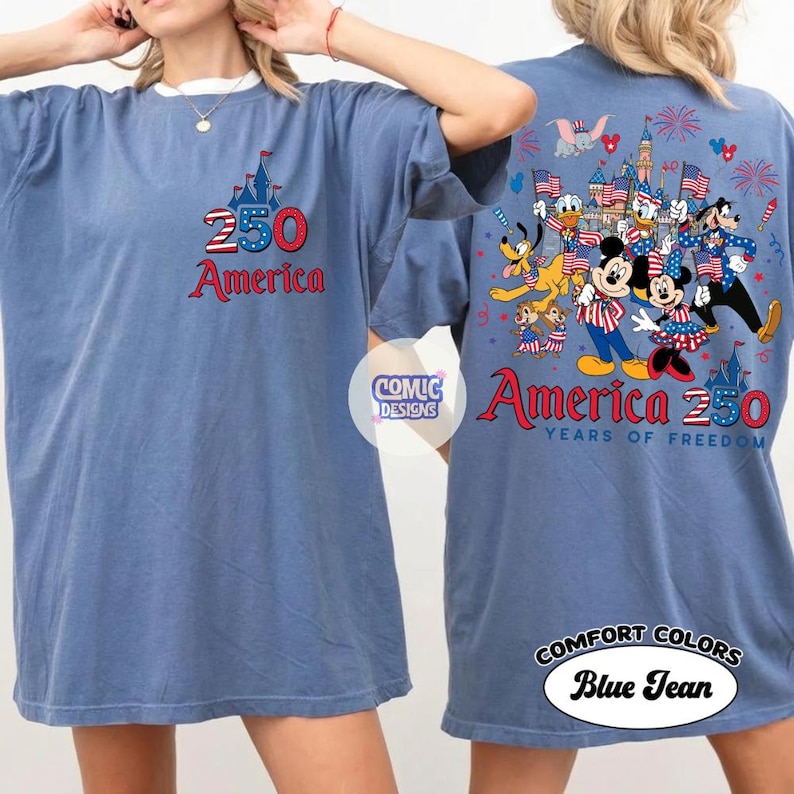 以下が含まれることがあります： フロントに城のグラフィックと「250 America」の文字が入ったブルーデニムのComfort Colors Tシャツ。背面には、旗と花火で祝うディズニーキャラクターのグラフィックと「America 250 Years of Freedom」の文字が描かれています。