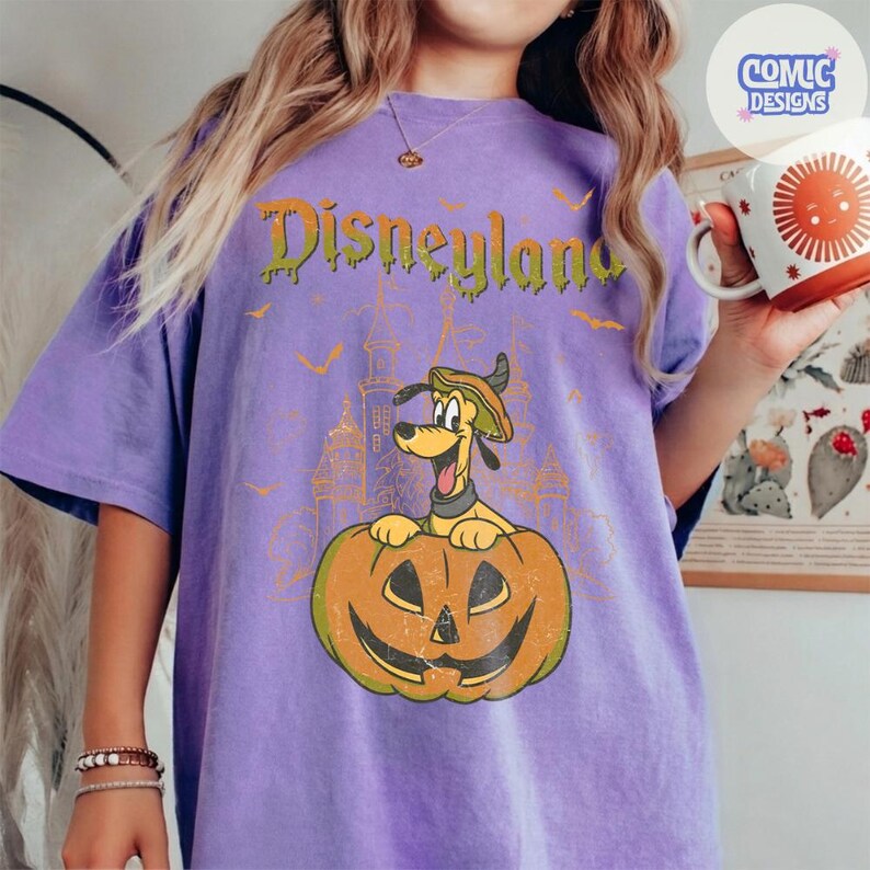 Może przedstawiać: Fioletowy t-shirt z kr&oacute;tkim rękawem z grafiką przedstawiającą Pluto w kapeluszu siedzącego w dyni. Nad grafiką znajduje się napis Disneyland wykonany zieloną, ściekającą czcionką. Wok&oacute;ł wzoru rozmieszczone są małe pomarańczowe nietoperze.