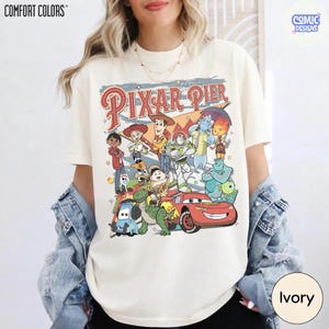 Pu&ograve; includere: T-shirt color avorio con una grafica dei personaggi di Pixar Pier. Il design include personaggi di Toy Story, Monsters, Inc. e Cars, con le parole "Pixar Pier" in stile vintage. La maglietta &egrave; del marchio Comfort Colors.