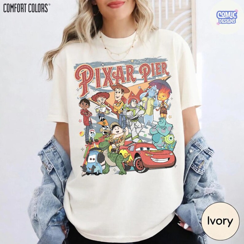 Pu&ograve; includere: T-shirt color avorio con una grafica dei personaggi di Pixar Pier. Il design include personaggi di Toy Story, Monsters, Inc. e Cars, con le parole "Pixar Pier" in stile vintage. La maglietta &egrave; del marchio Comfort Colors.