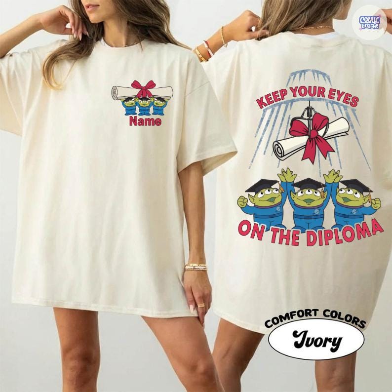 May include: Ivory-colored t-shirt featuring a cartoon design of three green aliens holding a diploma with a red bow. The front of the shirt has the word "Name" below the aliens. The back of the shirt says "Keep Your Eyes On The Diploma".
