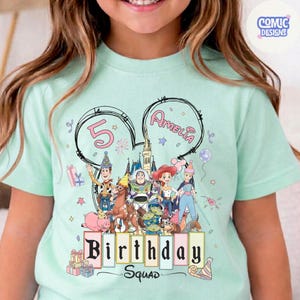 May include: A light green t-shirt featuring a colorful graphic of Disney's Toy Story characters. The design includes the text "Birthday Squad" and the name "Amelia" with the number "5". The shirt has short sleeves and a round neckline.