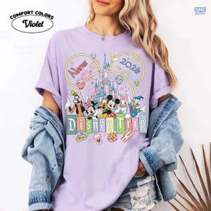 May include: A violet Comfort Colors t-shirt featuring a Disney Trip design. The design includes Disney characters, a castle, fireworks, and the text "Name" and "2020". The shirt has a crew neck and short sleeves.