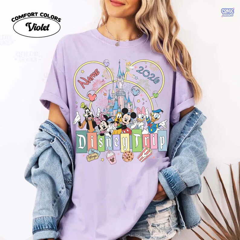 May include: A violet Comfort Colors t-shirt featuring a Disney Trip design. The design includes Disney characters, a castle, fireworks, and the text "Name" and "2020". The shirt has a crew neck and short sleeves.
