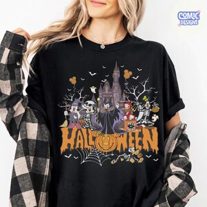 Puede incluir: Una camiseta negra de manga corta con un estampado gr&aacute;fico de personajes de dibujos animados con disfraces de Halloween frente a un castillo. El dise&ntilde;o incluye el texto "HALLOWEEN" en letras grandes de color naranja, una calabaza, telara&ntilde;as y murci&eacute;lagos.