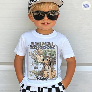 May include: White t-shirt featuring a cartoon illustration of Pluto from Disney's Animal Kingdom. The design includes the text "ANIMAL KINGDOM" and a space for a name. The shirt is worn by a child wearing sunglasses and a checkered hat.