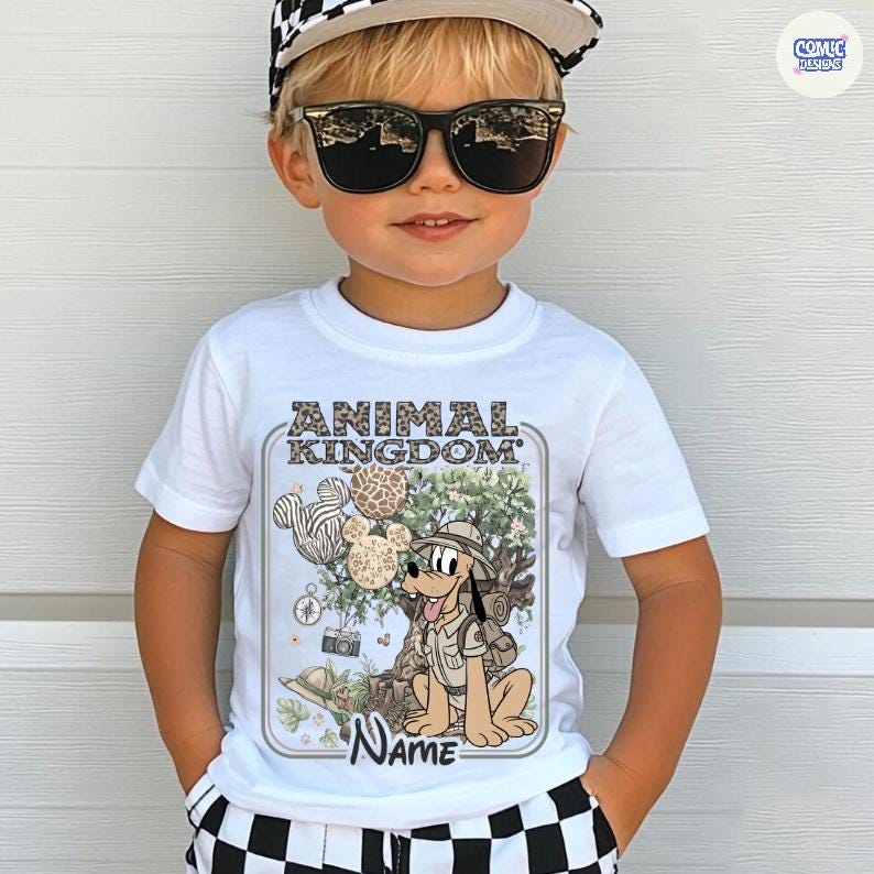 May include: White t-shirt featuring a cartoon illustration of Pluto from Disney's Animal Kingdom. The design includes the text "ANIMAL KINGDOM" and a space for a name. The shirt is worn by a child wearing sunglasses and a checkered hat.