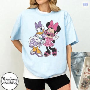 Puede incluir: Una camiseta azul claro con un estampado gr&aacute;fico de Daisy Duck y Minnie Mouse. Daisy lleva un vestido y un lazo con estampado floral morado, mientras que Minnie lleva un vestido y un lazo de lunares rosas con gafas de sol rosas.