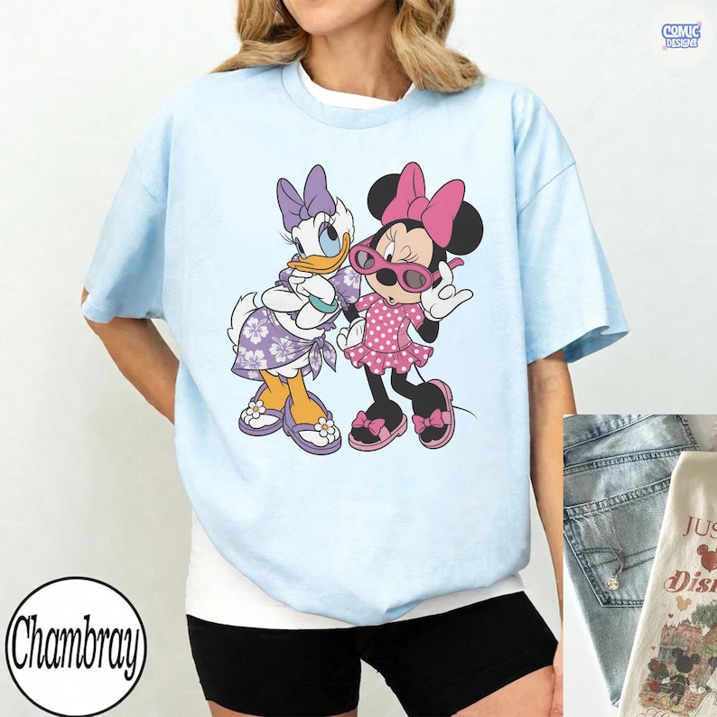 Puede incluir: Una camiseta azul claro con un estampado gr&aacute;fico de Daisy Duck y Minnie Mouse. Daisy lleva un vestido y un lazo con estampado floral morado, mientras que Minnie lleva un vestido y un lazo de lunares rosas con gafas de sol rosas.