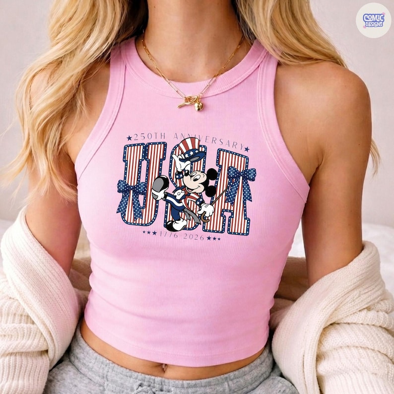 Op de afbeelding: Een lichtroze geribbelde tanktop met een afbeelding van Mickey Mouse in een patriottische hoed en outfit. De tekst luidt 250TH ANNIVERSARY USA 1776 2026, met rood-wit gestreepte letters en blauwe strikken met witte sterren aan de zijkanten.
