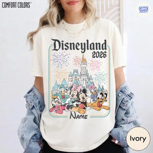 Op de afbeelding: Ivoor kleurig T-shirt met een Disneyland 2026-afbeelding met cartoonfiguren, een kasteel en vuurwerk. Het ontwerp bevat het woord "Name" onder de karakters. Het shirt heeft het "Comfort Colors" logo.