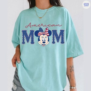 May include: A light blue t-shirt featuring the text "American MOM" in a patriotic design. The word "MOM" is stylized with a Minnie Mouse graphic, incorporating a red, white, and blue color scheme. The shirt has a relaxed fit.