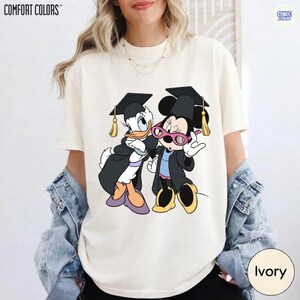 Puede incluir: Camiseta Comfort Colors color marfil con un gr&aacute;fico de Donald Duck y Minnie Mouse con atuendo de graduaci&oacute;n. Los personajes llevan birretes y togas negras. La camiseta tiene la palabra "COMFORT COLORS" impresa en la parte superior.