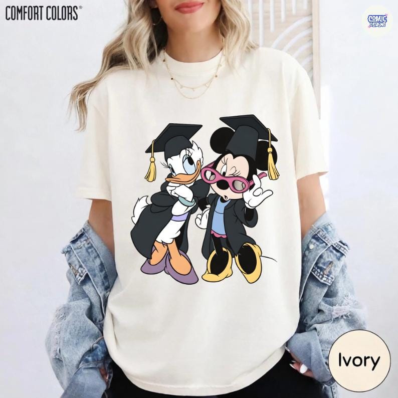 Puede incluir: Camiseta Comfort Colors color marfil con un gr&aacute;fico de Donald Duck y Minnie Mouse con atuendo de graduaci&oacute;n. Los personajes llevan birretes y togas negras. La camiseta tiene la palabra "COMFORT COLORS" impresa en la parte superior.