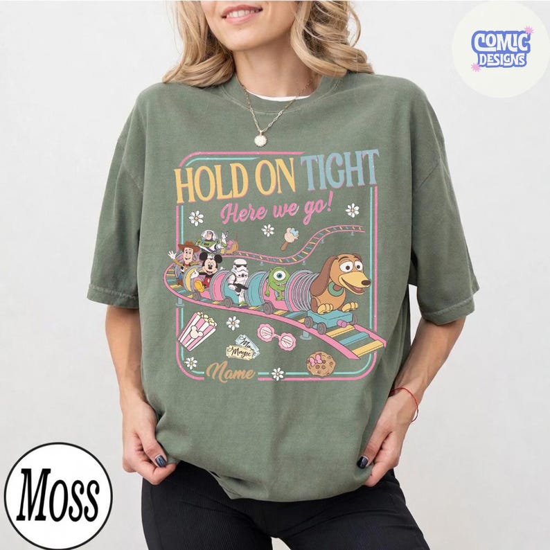 May include: Olive green t-shirt with a colorful graphic featuring cartoon characters on a roller coaster with the text "HOLD ON TIGHT Here we go!" in a retro style. The shirt has a relaxed fit.