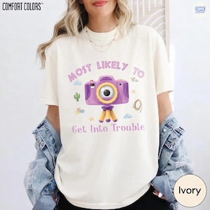 May include: An ivory short-sleeve t-shirt featuring a purple camera graphic on a tripod. The text reads "MOST LIKELY TO Get Into Trouble" in purple letters. Small colorful icons including a cactus, clouds, stars, and a rope surround the camera.