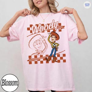 May include: A light pink t-shirt featuring a graphic of the character Woody from Toy Story. The design includes the text Woody in a stylized font, a checkered pattern, and a line drawing of the character alongside a full-color illustration.