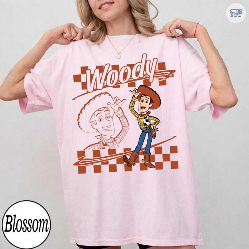May include: A light pink t-shirt featuring a graphic of the character Woody from Toy Story. The design includes the text Woody in a stylized font, a checkered pattern, and a line drawing of the character alongside a full-color illustration.