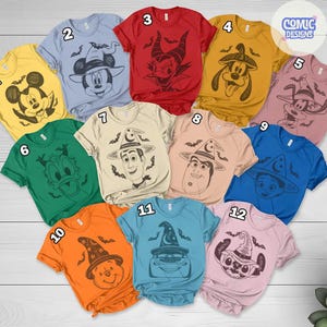 May include: A collection of colorful t-shirts featuring various cartoon characters in Halloween-themed designs. The shirts display characters like Mickey Mouse, Woody, and Winnie the Pooh, each wearing witch hats and surrounded by bats. The shirts are in various colors.
