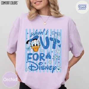 May include: Orchid-colored t-shirt with a graphic that says "school's out for Disney" in blue. The design includes Donald Duck, popcorn, a castle, and other Disney-themed elements. The shirt is from Comfort Colors.