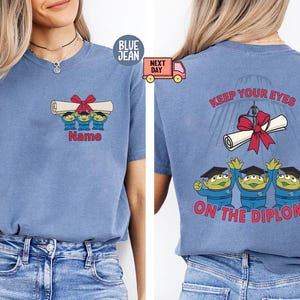 May include: Blue short-sleeved t-shirt with a graduation-themed graphic. The front features three green aliens holding a diploma with a red bow, with the text "Name" below. The back says "Keep Your Eyes On The Diploma".