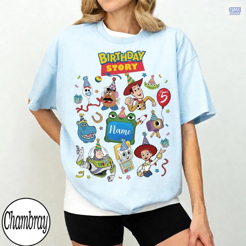 May include: A light blue t-shirt featuring a colorful graphic of various animated toy characters wearing party hats. The text "BIRTHDAY STORY" appears at the top, and a green rectangular frame in the center includes the word "Name" in script.