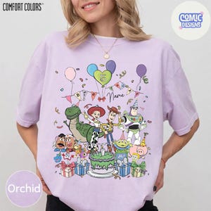 Puede incluir: Una camiseta de color morado claro con un estampado gr&aacute;fico de varios personajes de juguetes animados rodeando un pastel de cumplea&ntilde;os. El dise&ntilde;o incluye globos, una pancarta con el texto 'Birthday Name' y el n&uacute;mero 5 dentro de un coraz&oacute;n. La camiseta tiene mangas cortas y cuello redondo.