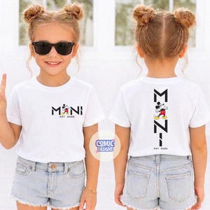 May include: White t-shirts with a Mickey Mouse design. The front of the shirt says "MINI" with Mickey Mouse and "EST. 2026". The back of the shirt has a vertical design with Mickey Mouse and "EST. 2026". The shirts are worn by a young girl.