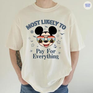 May include: Cream-colored t-shirt featuring a graphic of Mickey Mouse wearing sunglasses with a sunset design. The text "MOST LIKELY TO Pay For Everything" is printed above and below the character in navy blue.