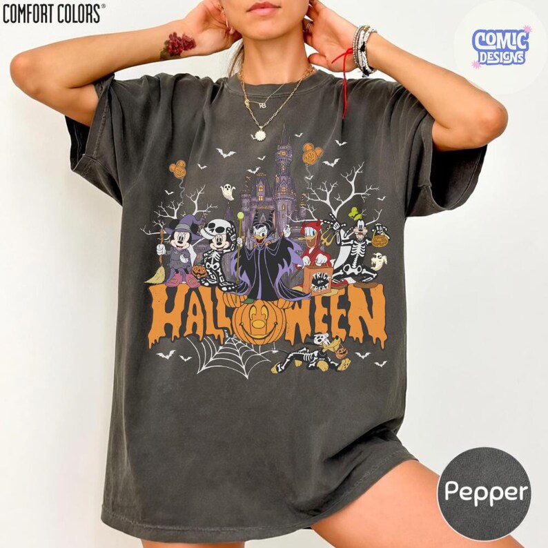 Puede incluir: Una camiseta de manga corta gris carb&oacute;n con un estampado gr&aacute;fico de varios personajes de dibujos animados con disfraces de Halloween, incluyendo esqueletos y una bruja, con una calabaza naranja y el texto HALLOWEEN en el frente.
