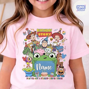 May include: A light pink t-shirt featuring a colorful graphic of Toy Story characters including Woody, Buzz Lightyear, and Jessie. The design includes the text Birthday Story and You've Got A Friend Who's Three, with a green frog frame for a name.