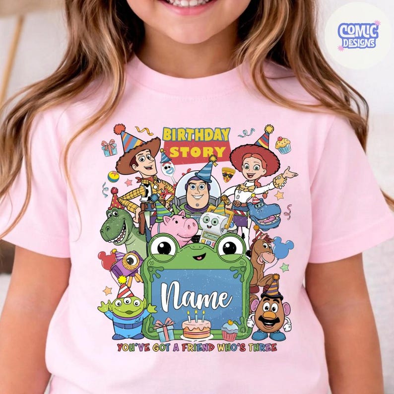 May include: A light pink t-shirt featuring a colorful graphic of Toy Story characters including Woody, Buzz Lightyear, and Jessie. The design includes the text Birthday Story and You've Got A Friend Who's Three, with a green frog frame for a name.
