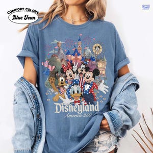 Puede incluir: Una camiseta de color azul vaquero con un gr&aacute;fico de Mickey Mouse, Minnie Mouse, el Pato Donald, Goofy y Pluto vistiendo ropa patri&oacute;tica roja, blanca y azul. El dise&ntilde;o incluye un castillo, fuegos artificiales y el texto Disneyland America 250.