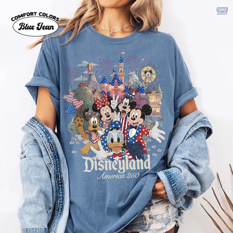 Puede incluir: Una camiseta de color azul vaquero con un gr&aacute;fico de Mickey Mouse, Minnie Mouse, el Pato Donald, Goofy y Pluto vistiendo ropa patri&oacute;tica roja, blanca y azul. El dise&ntilde;o incluye un castillo, fuegos artificiales y el texto Disneyland America 250.